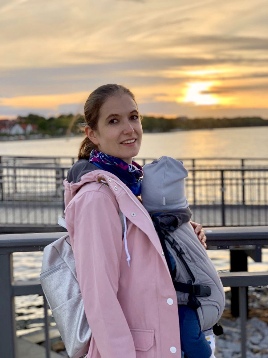 Mama und Sohn mit der Babytrage im Urlaub unterwegs. Erster Urlaub mit Baby am Meer beim Sonnenuntergang.