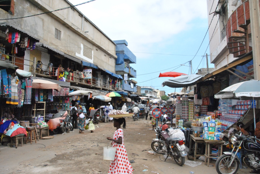 Vue d’une petite rue du marché où se mêlent magasins formels, stands sur les trottoirs et vendeurs ambulants.