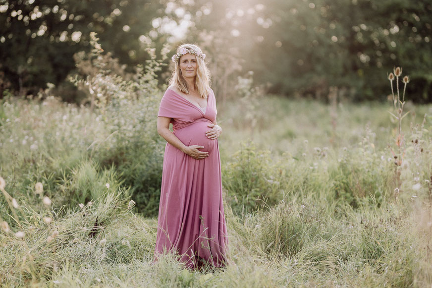 eine blonde, schwangere Frau in gelbem Kleid mit weissen Punkten steht vor einer Reihe Sonnenblumen und hält die Hände schützend an den Bauch