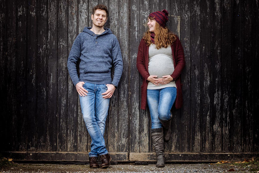 Fröhliches Paar steht mit dem Rücken an einem alten Holztor; sie hält ihre Hände an den Babybauch.