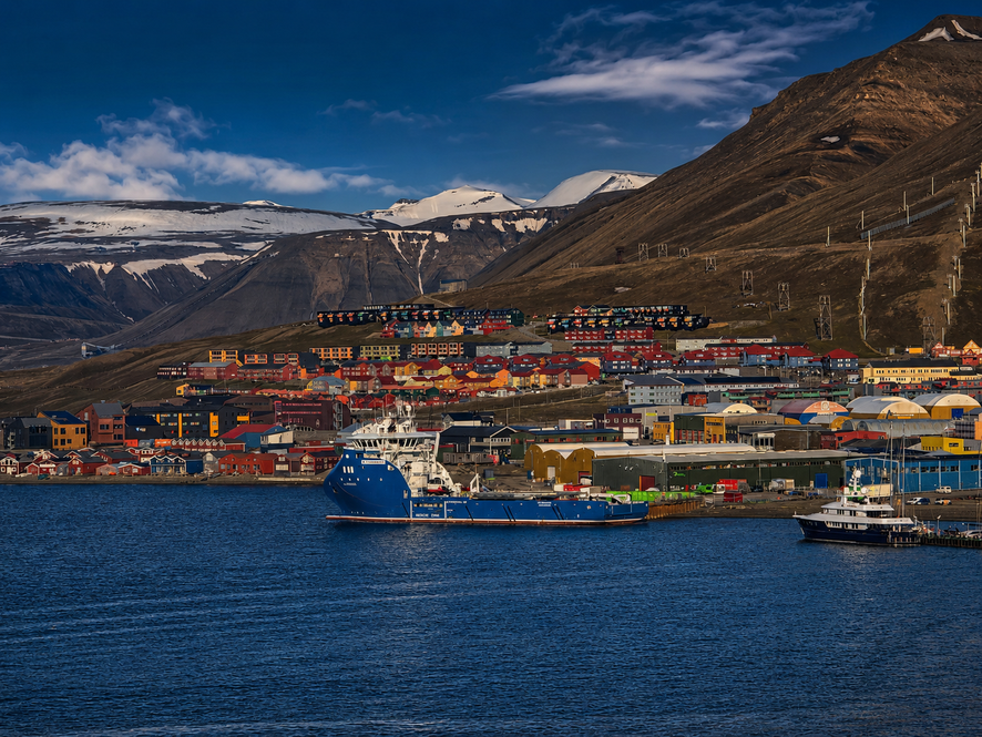 Panoramablick auf Longyearbyen auf Spitzbergen mit bunten Häusern, Hafen, Schiffen und schneebedeckten Bergen im Hintergrund.