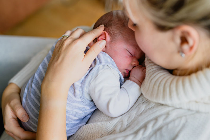Nahaufnahme einer Mutter, die ihr schlafendes Baby im Arm hält und liebevoll dessen Ohr streichelt.