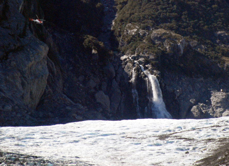 A helicopter dwarfed by the Chancellor Ridge coming in to land on the mid-tongue plateau of the Fox Glacier and 