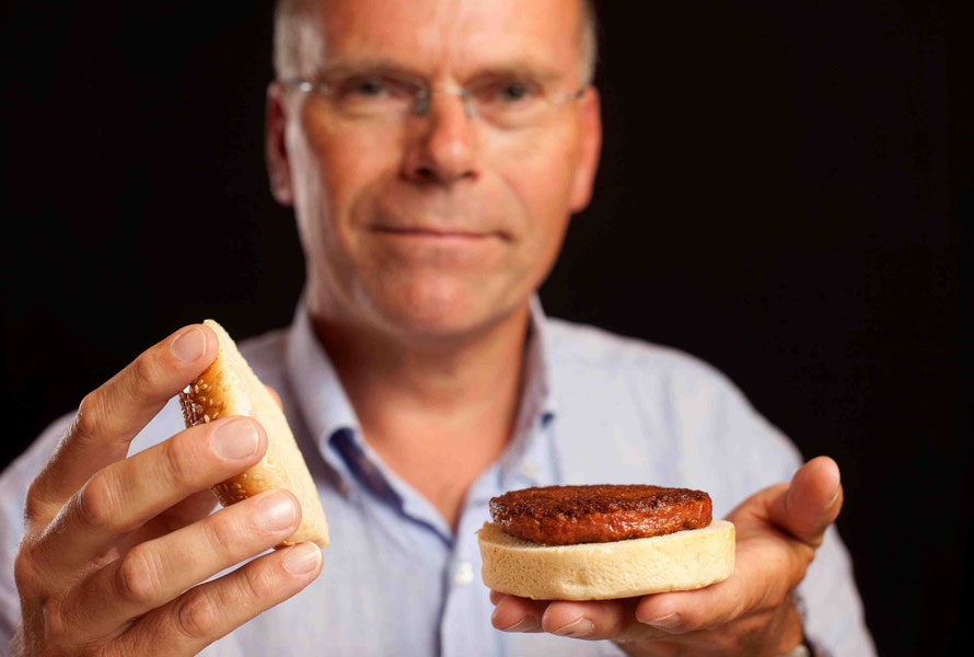Ein Mann mit Brille und Oberhemd hält ein aufgeklapptes Hamburgerbrötchen in die Kamera, das mit einem Fleisch-Patty aus Kulturfleisch belegt ist.