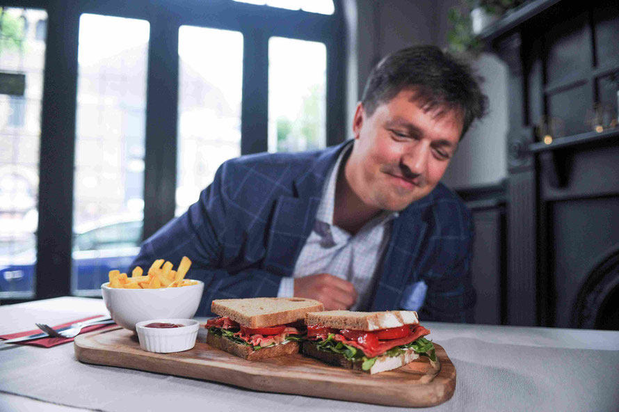 Ein Mann sitzt in einem Restaurant und beäugt lächelnd zwei Brotstullen, zwischen denen Salat und Speck hervorschaut.