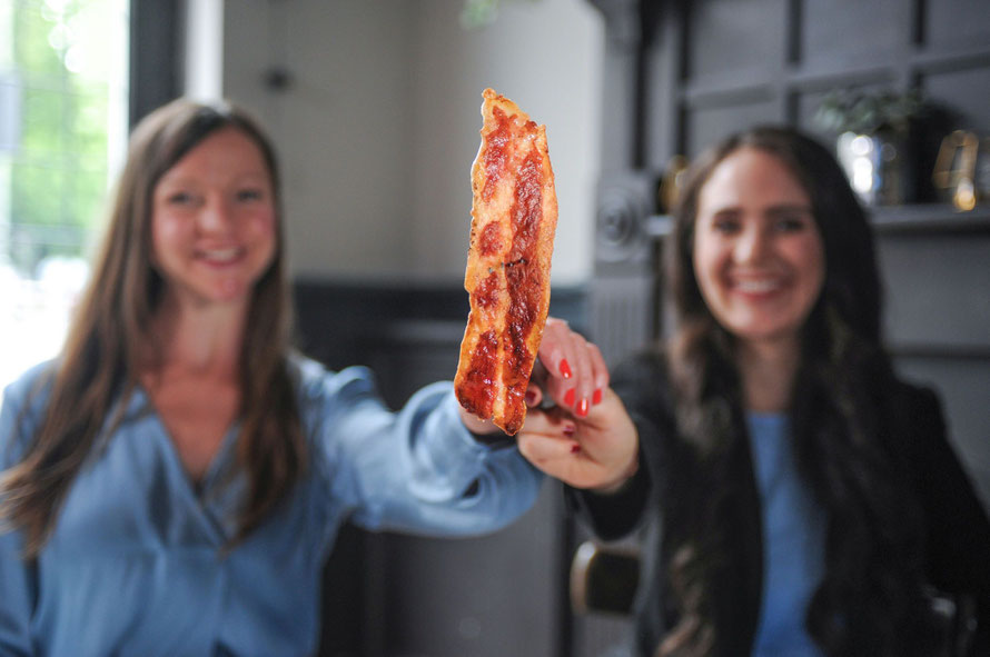 Zwei langhaarige Frauen halten ein Stück Bacon in die Kamera, das auf einer Gabel steckt.