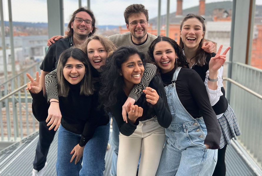 Sieben junge Erwachsene lachen in die Kamera. Einige haben die Arme um die Schultern der anderen gelegt, andere zeigen das Peace-Zeichen. Im Hintergrund sind die Dächer einer Stadt zu erkennen.