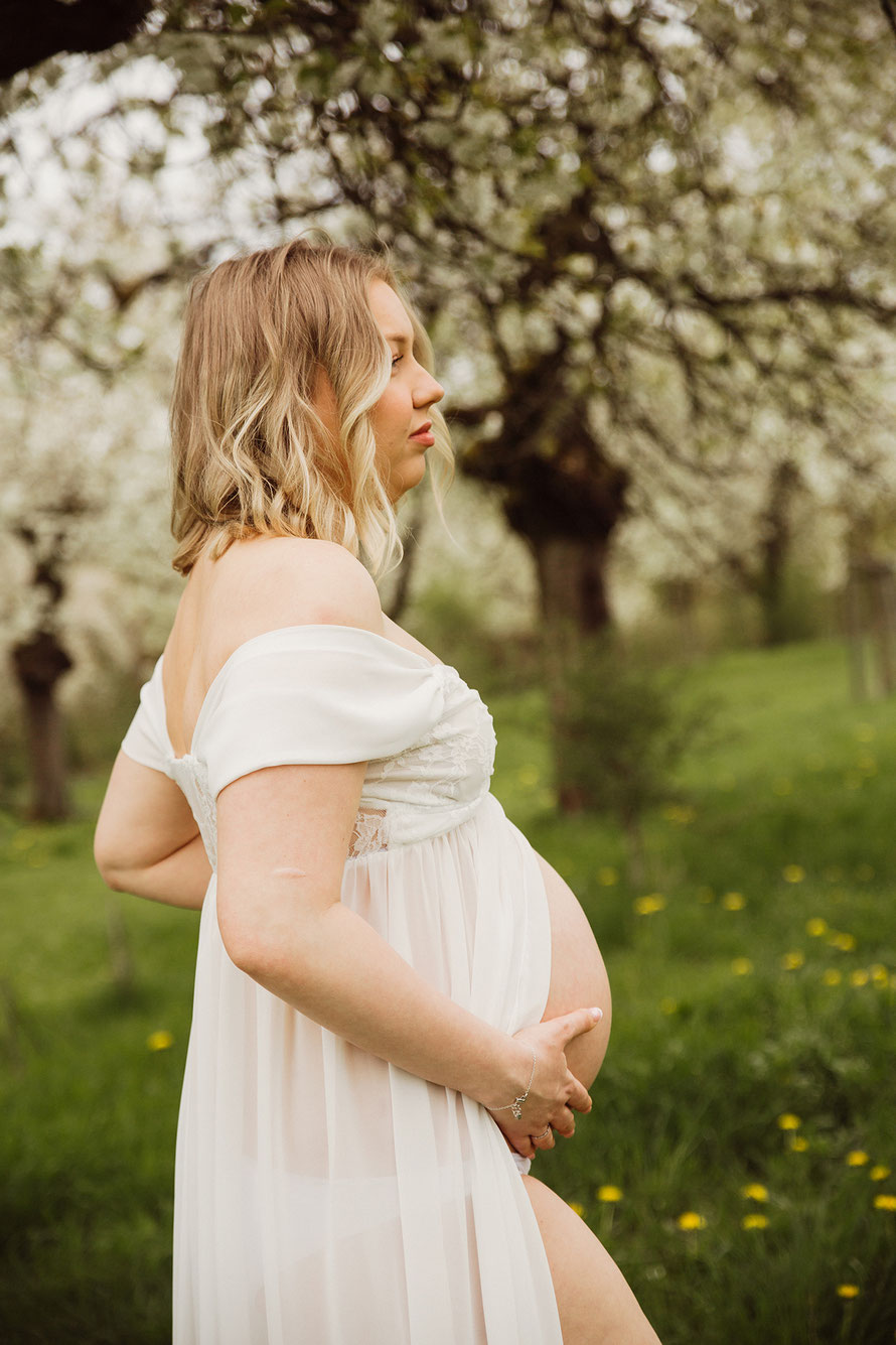 Schwangere Frau im weißen Kleid bei einem Babybauchshooting in der Kirschplantage Treffurt – emotionale Schwangerschaftsfotografie in natürlicher Frühlingskulisse.