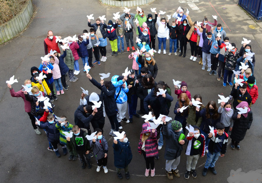 Das Bild zeigt die Schüler der Schule von oben. Sie stehen in Form eines Peace Zeichens und halten Tauben aus Papier hoch.