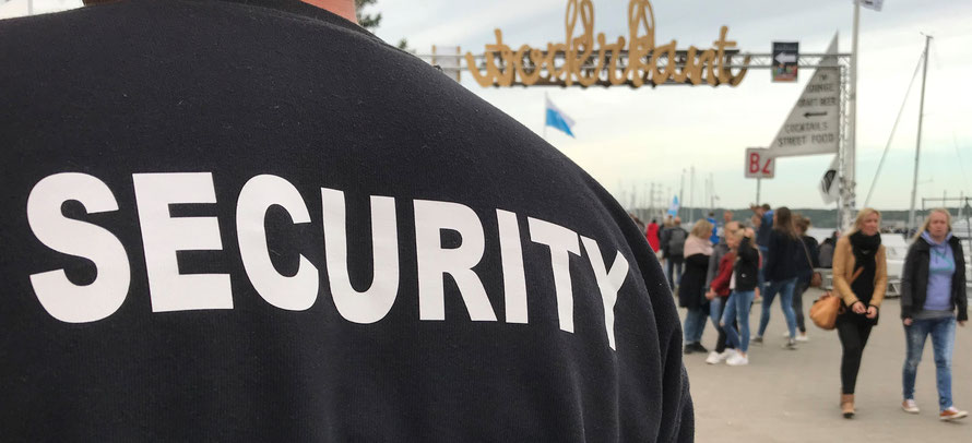 Rücken eines Sicherheitsmitarbeiters im schwarzen Shirt mit "SECURITY" in Großbuchstaben vor einem Durchgangstor bei der Kieler Woche. Auf dem Tor steht "Waterkant" und Menschen laufen oder schlendern im Hintergrund umher.  