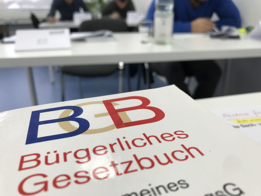 Im Vordergrung sieht man auf dem Dozebntentisch ein BGB (Bürgerliches Gesetzbuch), während im Hintergrund (verschwommen) 3 Schüler, hintereinander im Klassenraum sitzend, einen Test bearbeiten.