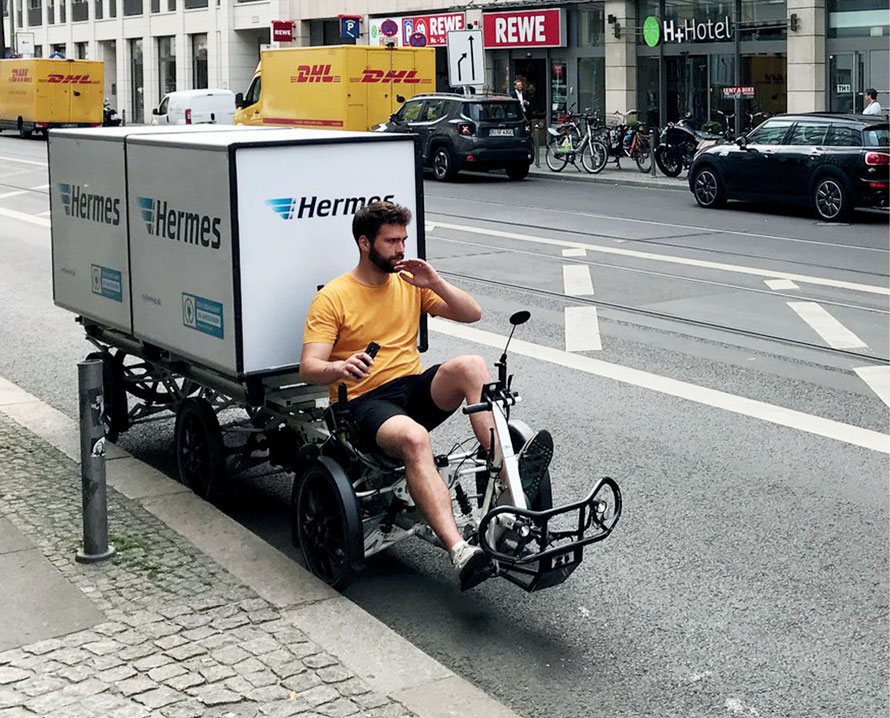 Hermeszusteller auf Lastenrad und konventionelle DHL-Zustellfahrzeuge im Hintergrund