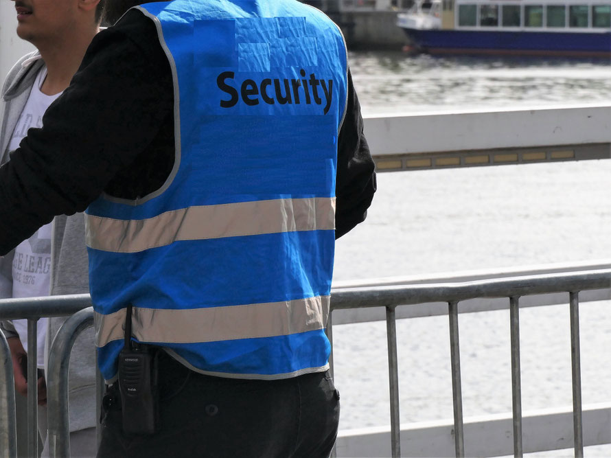 Sicherheits-Mitarbeiter mit weißem Schriftzug SECURITY auf dem Rücken einer blauen Warnweste vor einem Absperrgitter mit Menschen und Wasser dahinter (Hörnbrücke Kiel zur Kieler Woche)