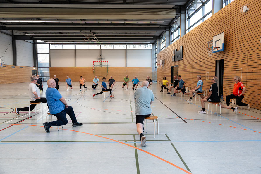 Gruppe von Teilnehmenden im Herz- und Rehasport trainiert gemeinsam mit Stühlen und gezielten Bewegungsübungen in der Sporthalle.