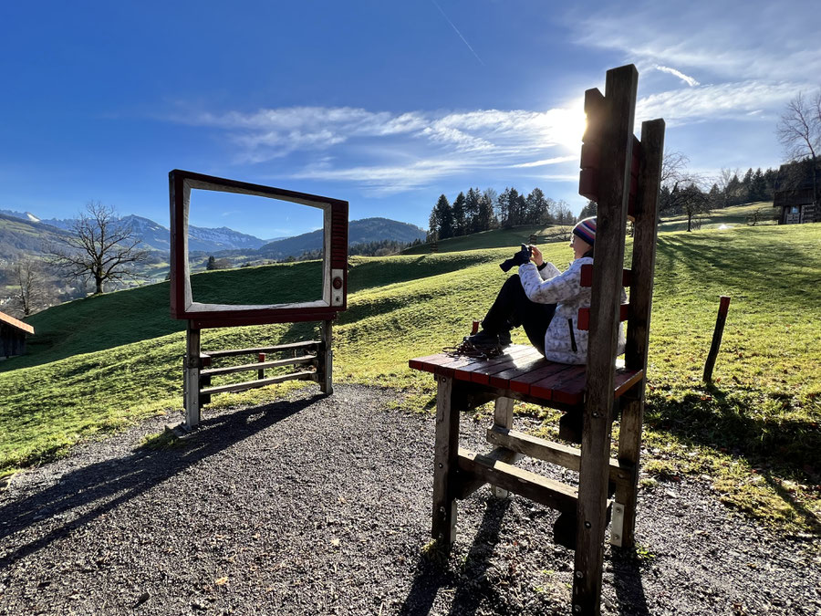 Ein Foto von Birgit Prinz in der Natur sitzend auf einem übergroßen Stuhl mit Blick auf einen Rahmen, der wie ein TV gestaltet ist