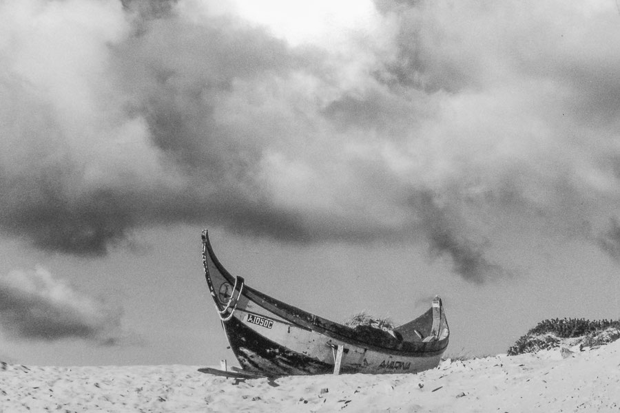 Ein großes Ruderboot liegt auf einer Düne im Sand. Darüber ein Himmel, an dem sich stürmische Wolken zusammenballen. Das Foto ist schwarzweiß.