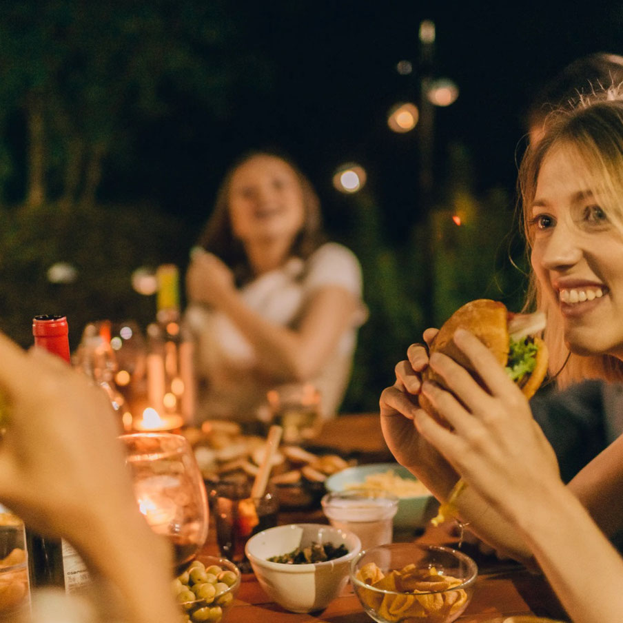 Zwei lachende Frauen und eine weitere Person sitzen an einem Esstisch mit Kerzenlicht und essen. 