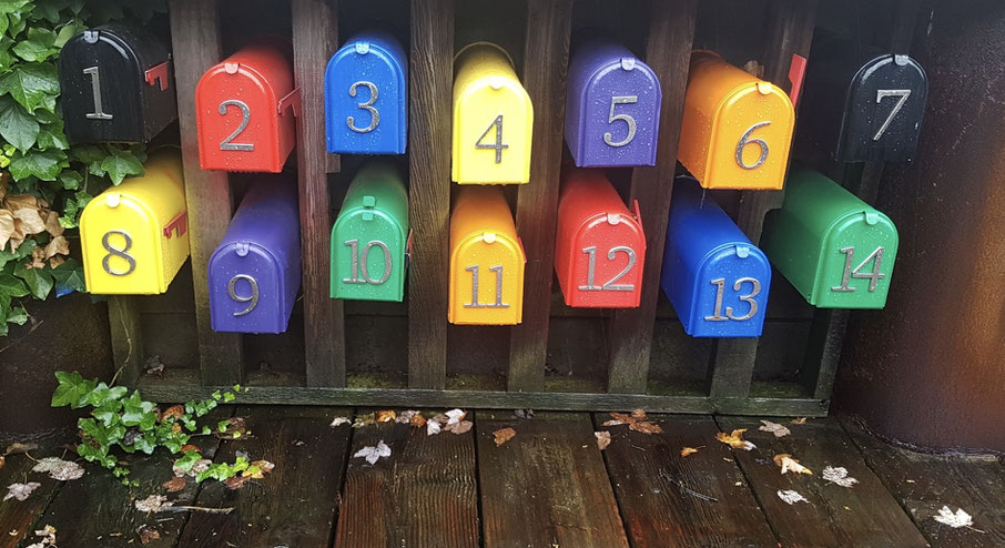Bunte, nummerierte Briefkästen, in zwei Reihen in einem Holzgestell übereinander angeordnet. Sie sind vom Regen nass und von 1 - 14 durchnummeriert.