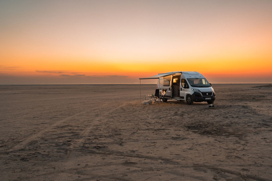 Vor einem Camper von Camperwelt Fischer genießt ein Mann mit Hund den Sonnenuntergang am Strand.