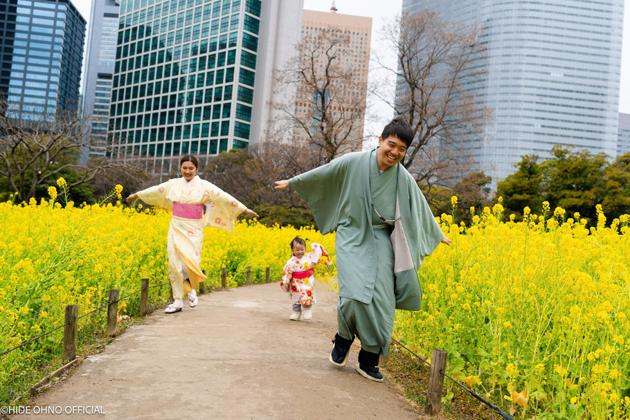 Singaporean family wearing kimono, playing joyfully among yellow rapeseed flowers in Hamarikyu Gardens, Tokyo, with skyscrapers in the background