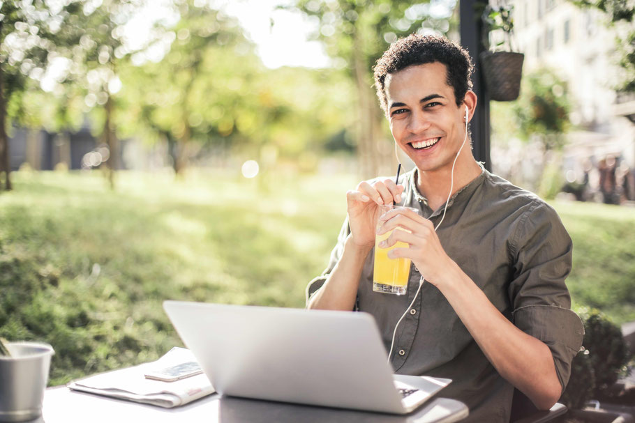 Ein junger Mann sitzt lächelnd irgendwo im Grünen hinter seinem Laptop, Knopf-Kopfhörer in den Ohren und ein orangefarbenes Getränk mit Strohhalm in den Händen.