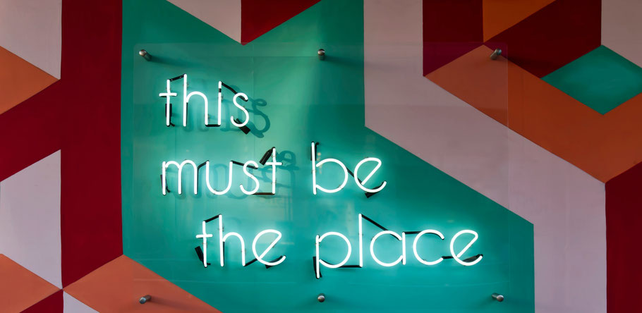 A wall filled with lilac, dark red and green areas. On the green area there are white neon letters hanging. The neon letters say: this must be the place.