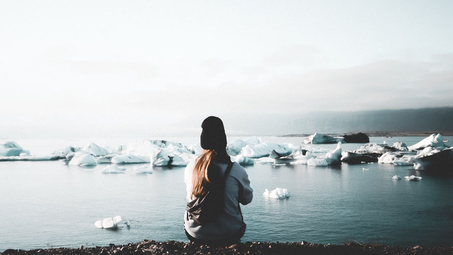 Eine junge Frau sitzt mit dem Rücken zum Betrachter an einem Gletschersee. Bild von Martin Balle auf Unsplash. 