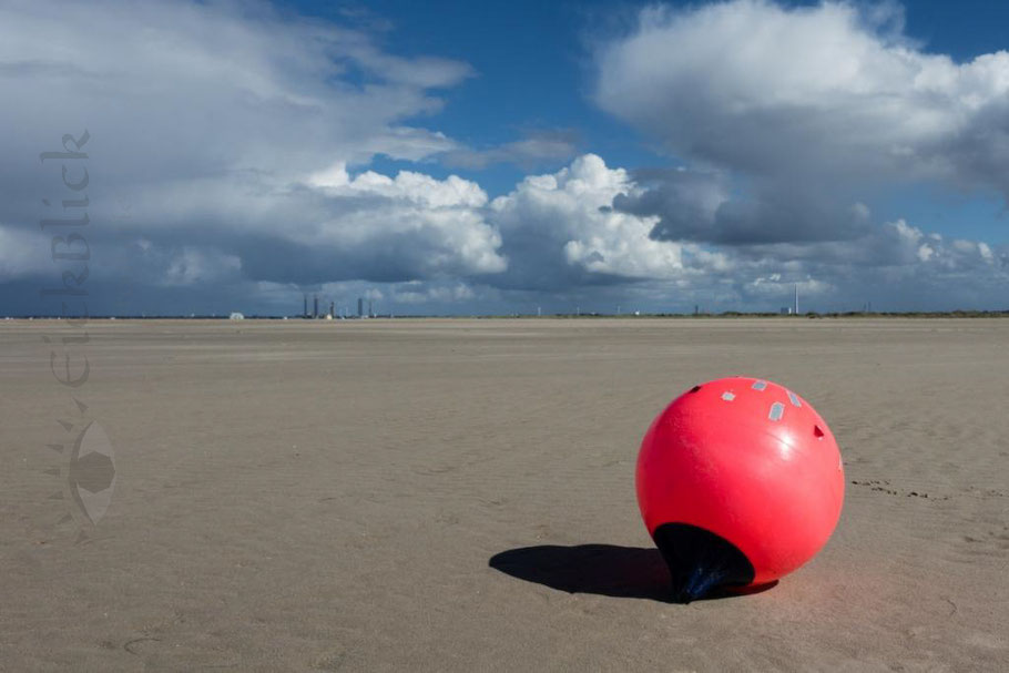 rote Boje als Strangut am weiten Strand von Fanö, im Hintergrund die Industrieanlagen von Esbjerg bei wolkigem Himmel 