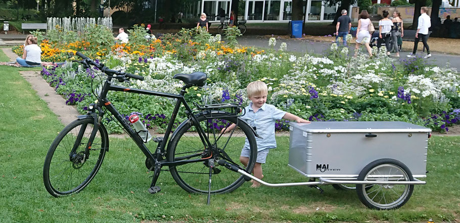 Ein kleiner, blonder Junge steht neben einem Gespann aus Fahrrad und dem Fahrradanhaenger Maiporter Z130 und schaut interessiert auf den Anhaenger