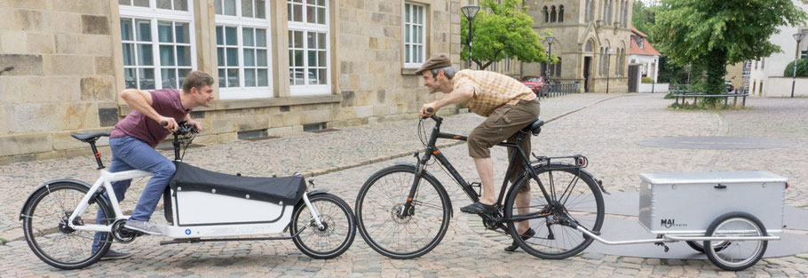 Lastenrad und Gespann aus Fahrrad und Lastenanhaenger stehen Vorderrad an Vorderrad voreinander und tun so, als wollten sie sich gegenseitig wegschieben