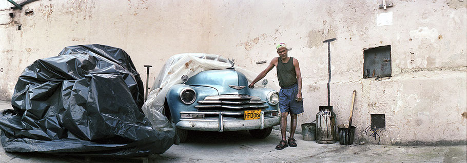 Mann zeigt blauen Oldtimer im Hinterhof in der Altstadt von Havanna als Farbphoto im Panoramaformat, Cuba