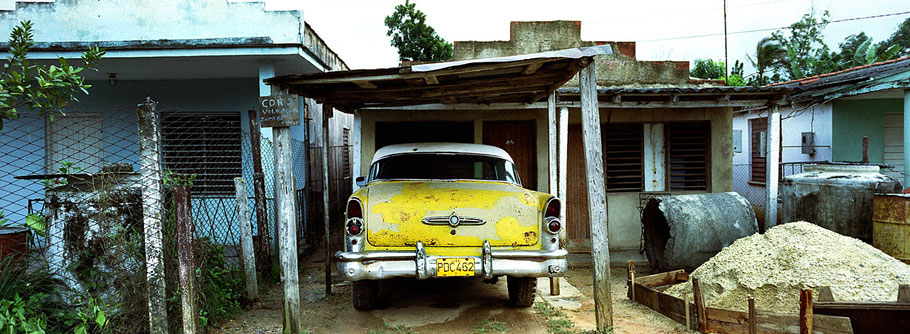 Gelber Oldtimer  steht vor einem Haus in der Zona 86  als Farbphoto im Panoramaformat, Cuba