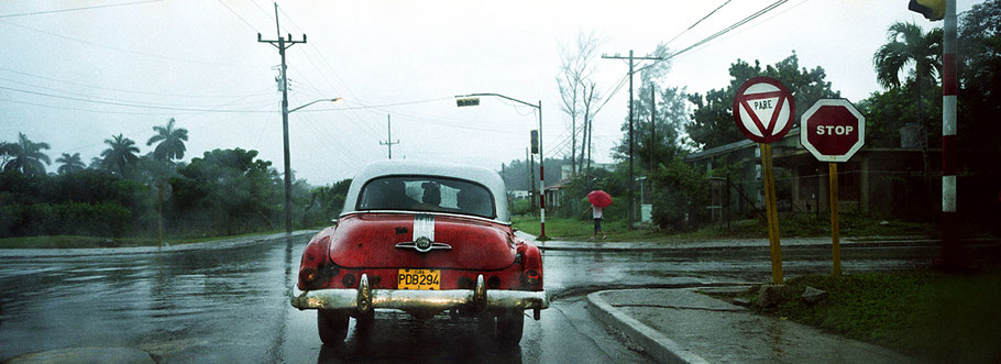 Roter Oldtimer steht im Regen am Stoppschild in Pinar del Rio als Farbphoto im Panoramaformat, Cuba