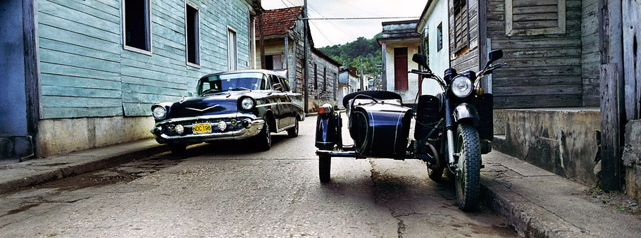 Schwarzer Oldtimer und Motorrad auf der Straße in Baracoa als Farbphoto im Panoramaformat, Cuba