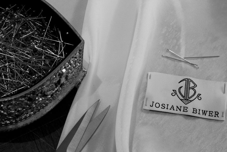 Schwarz Weiss Bild: Hintergrund: eine Schere, eine herzförmige, metallische Schatulle, gefüllt mit Stecknadeln. Im Vordergrund: ein Ausschnitt eines Stoffteiles auf welchem das Logo von Josiane Biwer gesteckt ist