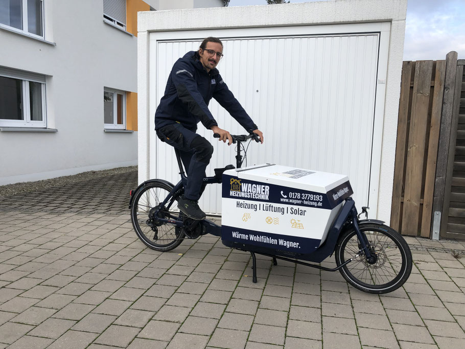 Heizungsbaumeister sitz auf einem Lastenrad. Das Lastenrad hat vor dem Lenker eine Werkzeug-Transportkiste mit Werbeaufdruck für die Firma Wagner Heizungstechnik.