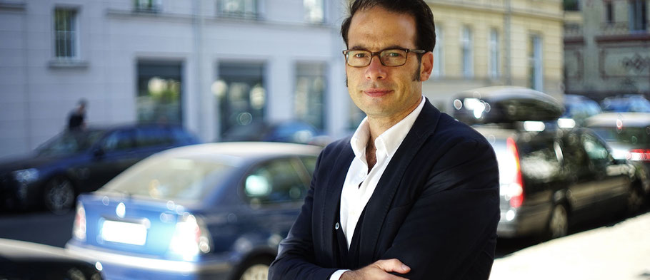 Wir sehen einen Rechtsanwalt vor einer Autoreihe stehen er hat eine Brille auf.