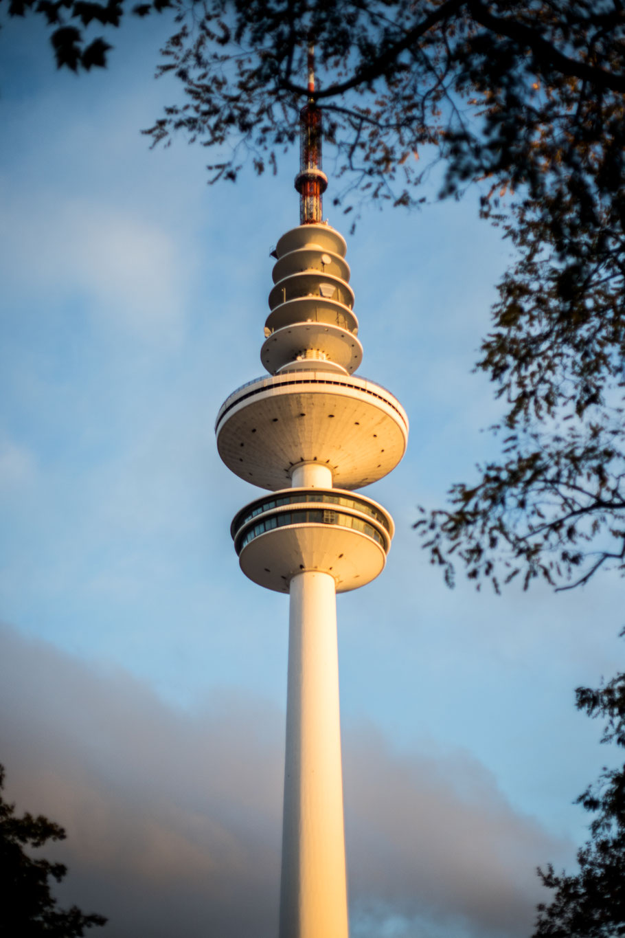 The Hamburg television tower against a partially cloudy autumn sky at sunset, surrounded by tree branches.