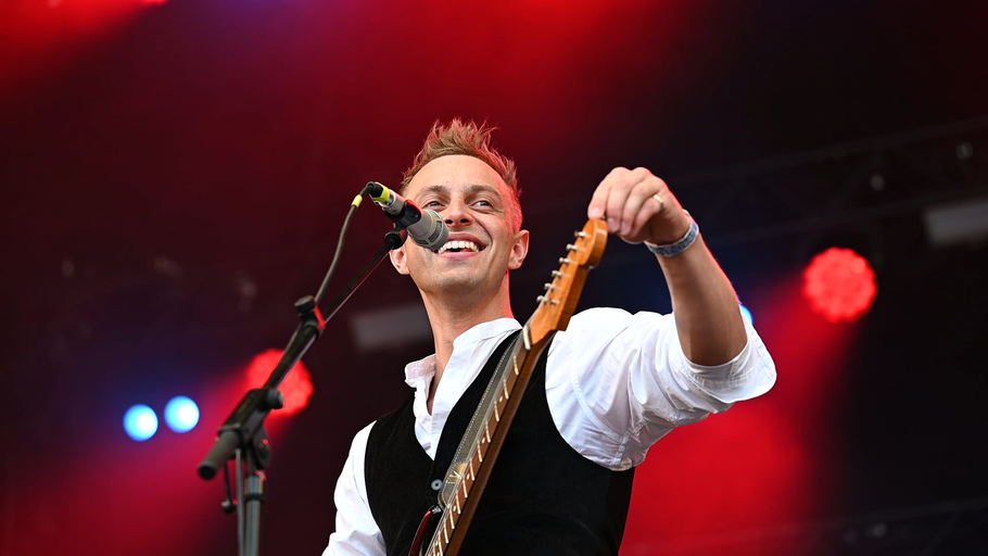 Der charismatischer Musiker Moritz Kruit steht auf einer großen Bühne, lächelt und hält eine Gitarre in der Hand, während er vor einem Mikrofon posiert.