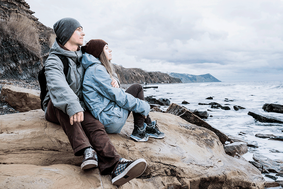Pärchen in Regenjacken sitzt chillig auf Felsen am Meer und schaut auf das Wasser.