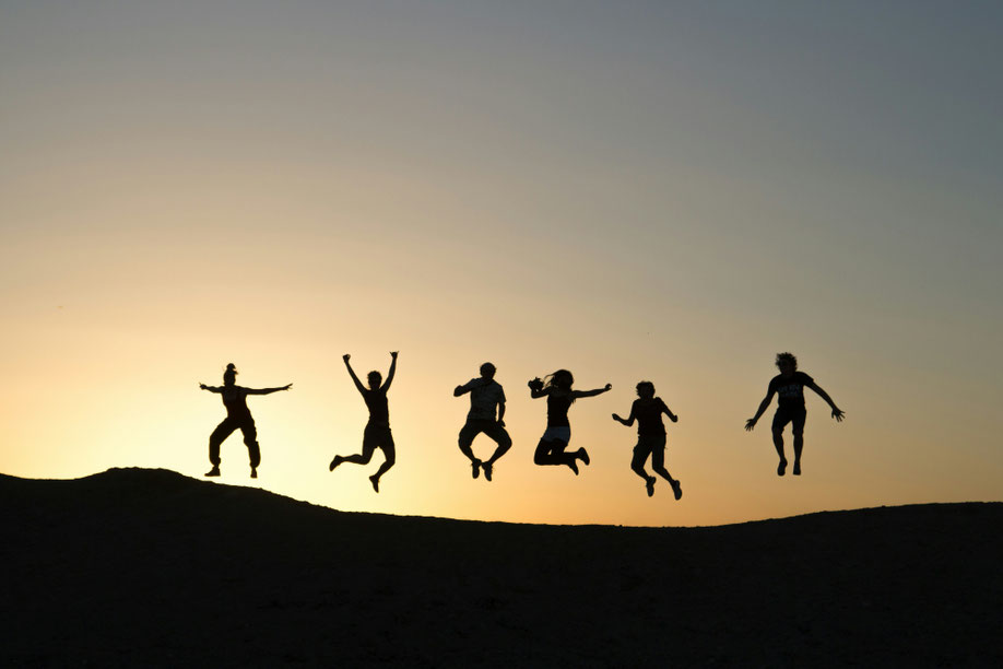 Auf der Kuppe eines Hügels sieht man sechs Menschen, die alle gleichzeitig in die Luft springen. Das Bild ist im goldenen Gegenlicht aufgenommen, die Figuren wirken wir bei einem Scherenschnitt.