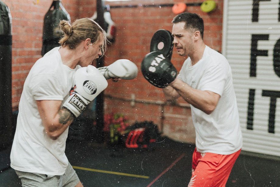 Zwei Männer trainieren Boxen mit Schlagkissen in einem Sportraum.