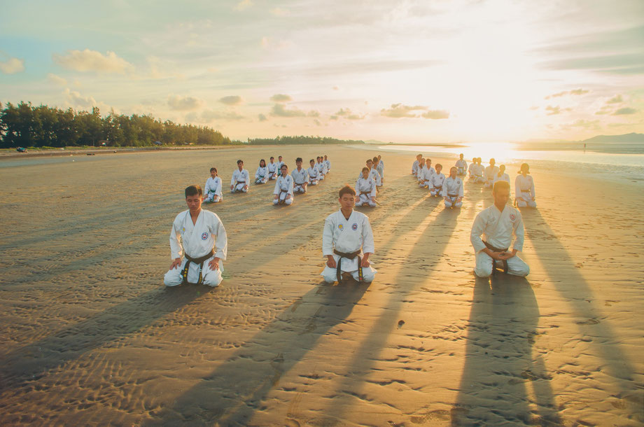 Karate-Meister grüßen gemeinsam an und meditieren vor dem Training am Strand. Im Hintergrund geht die Sonne auf.