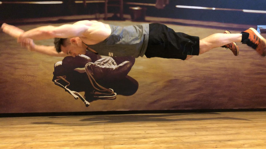 Ein Mann trainiert Liegestütze im Fitnessstudio. Er springt dabei hoch in die Luft und streckt alle vier Gliedmaßen von sich.