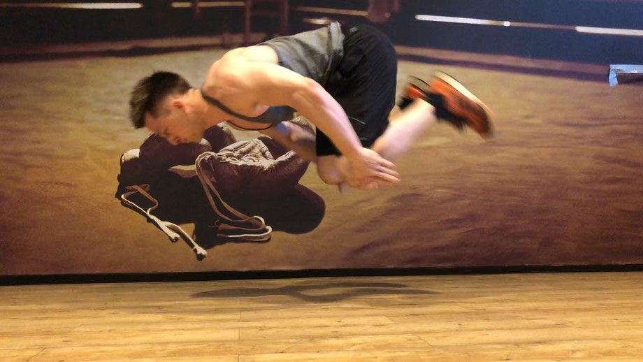 Ein Mann trainiert Liegestütze im Fitnessstudio. Er springt dabei hoch in die Luft und macht sich in der Luft klein, damit er mit den Händen seine Knie berühren kann.