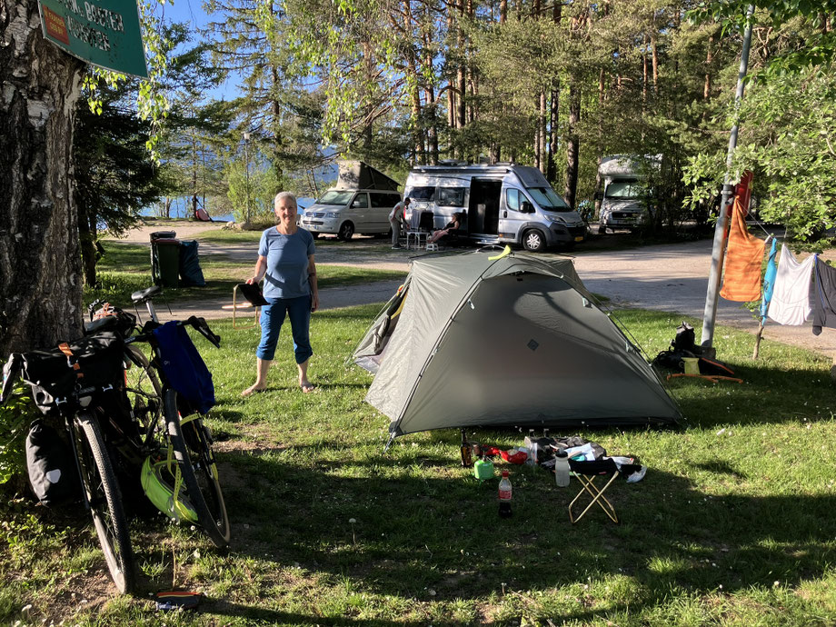 Bikepacking-Setup mit Gravelbike und Zelt auf einem Campingplatz am Faaker See in Österreich. Im Hintergrund stehen Wohnmobile und hohe Bäume direkt am Seeufer bei sonnigem Wetter.