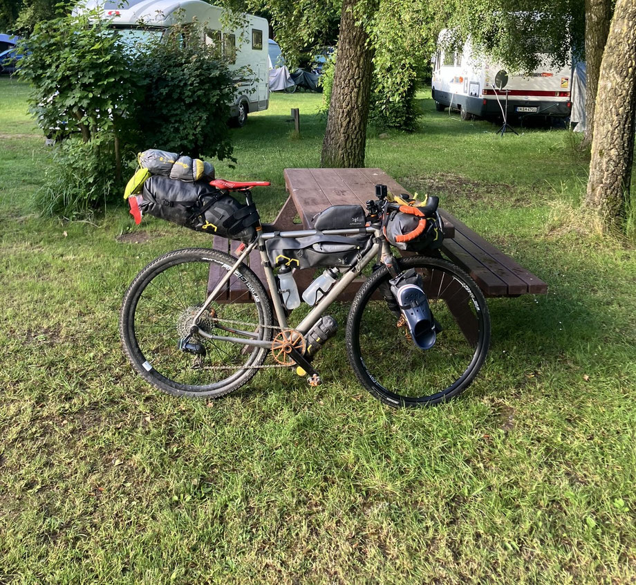 Bikepacking-Tour durch Kärnten – Campingplatz in Österreich – voll ausgestattetes Gravelbike mit Bikepacking-Taschen und Schlafsetup neben Holztisch auf grüner Wiese.