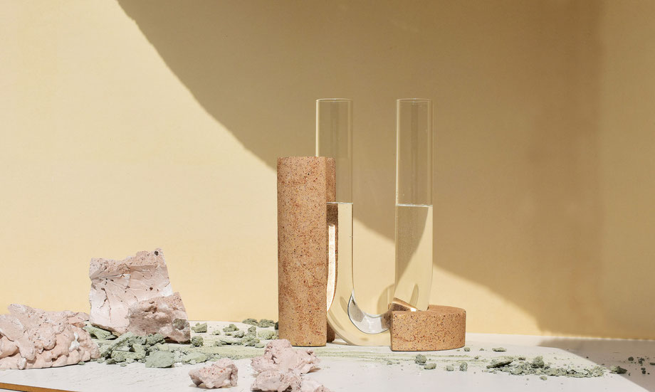Curved glass vase interlocked with two hand-sculpted natural stone bases, part of the Cochlea collection by COKI. Displayed on a white surface with scattered mineral fragments and soft yellow background lighting.