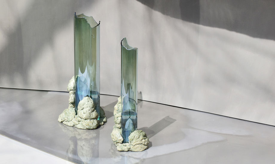 Two tall sculptural vases in flame-worked glass with jagged edges, anchored in hand-sculpted natural stone bases. Displayed on a smooth reflective surface with soft shadows, part of the Catharsis collection by COKI.