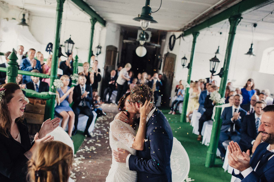 Die Brautleute küssen sich. Von oben fällt Konfetti. Die Gäste sind seitlich von ihnen, amüsieren sich und applaudieren